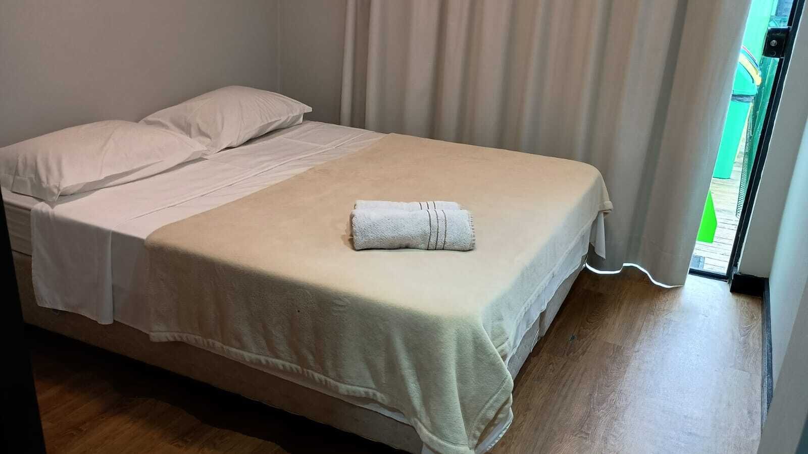 A neatly made bed with a white duvet and a folded towel on top, a wooden floor, and a view of a green outdoor area through a doorway.
