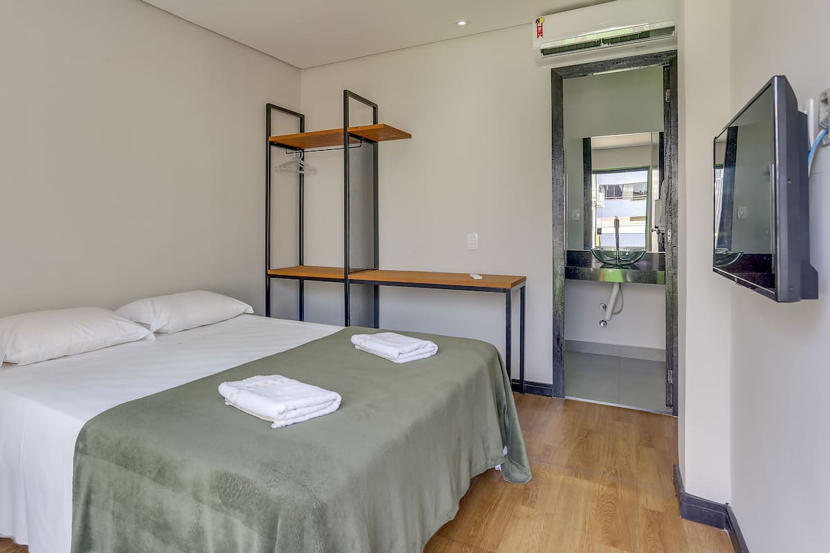 Une chambre d’hôtel avec un lit, une petite table, un miroir, une télévision et une salle de bain visible à travers une porte ouverte.