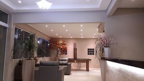 A modern living room with a sofa, a coffee table, and a dining table. There are potted plants and a large window. - Pousada Cantinho Oriental (Bertioga)