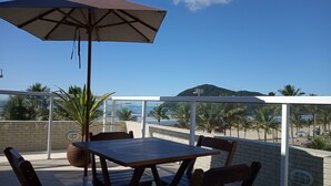 A balcony with a table and chairs, a large umbrella, and a view of the beach and mountains. - Pousada Cantinho Oriental (Bertioga)