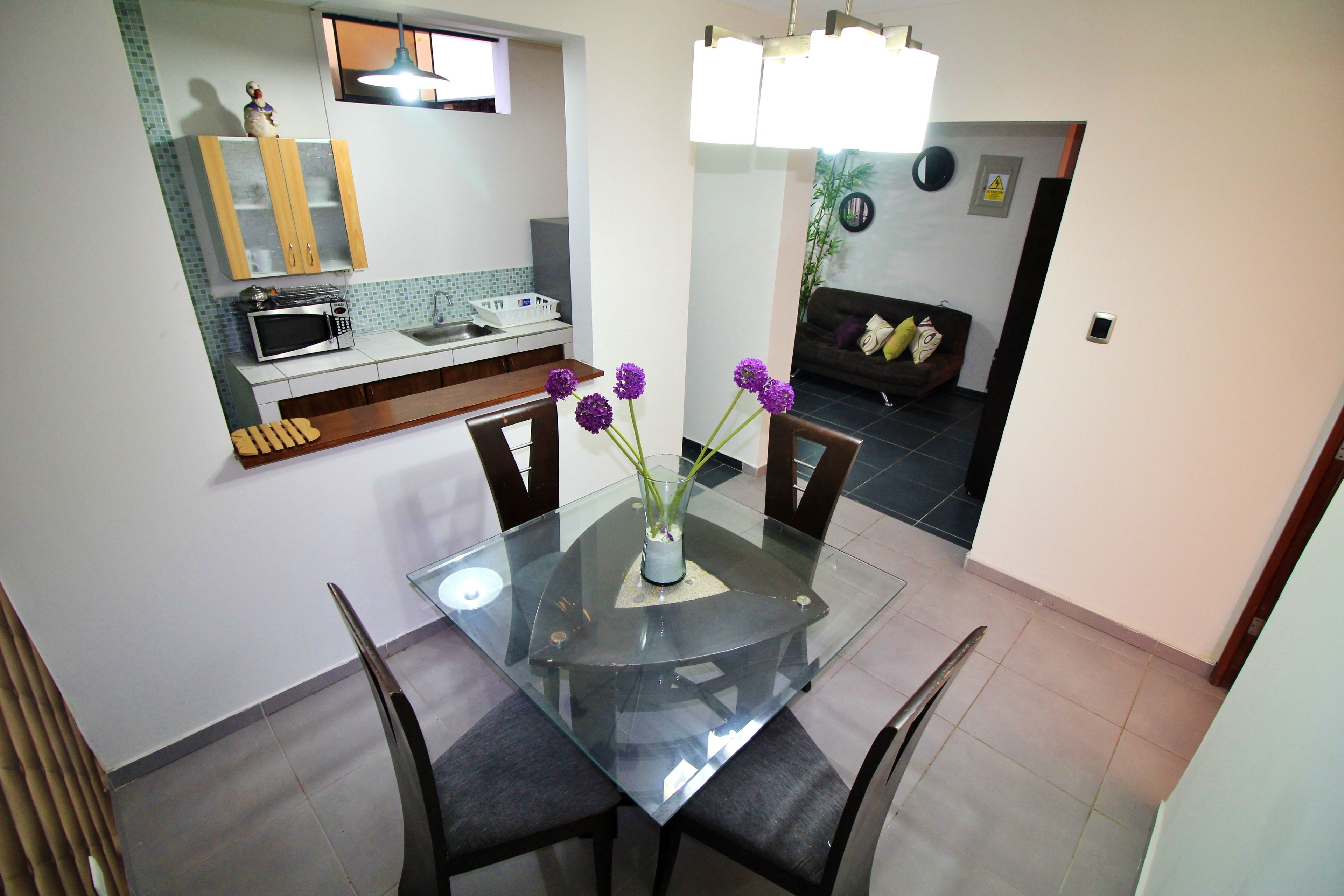 A modern kitchen with a glass dining table, chairs, and a vase with purple flowers.
