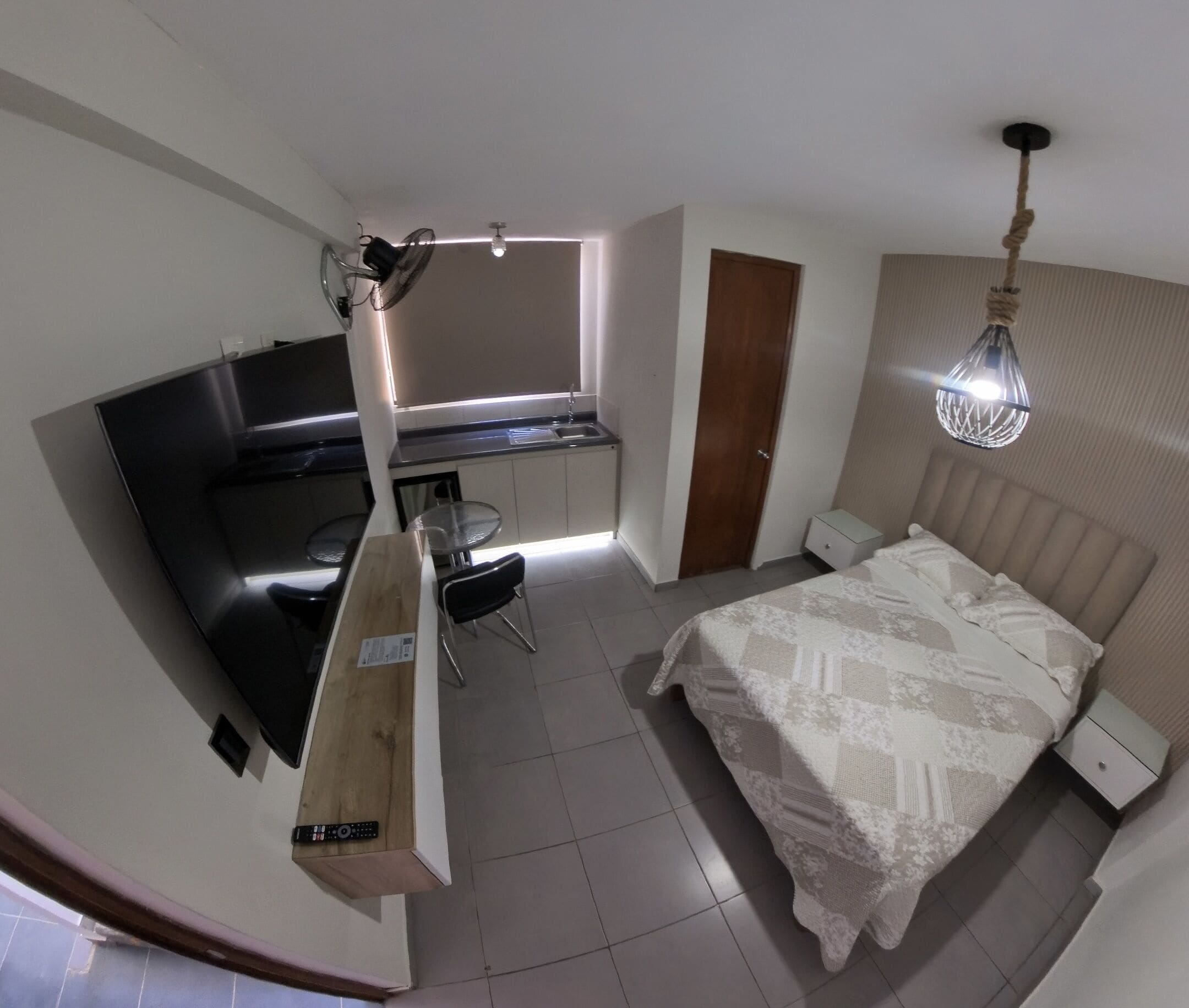 Un dormitorio compacto con cama, mesitas de noche, un escritorio con silla, una kitchenette pequeña y una ventana.