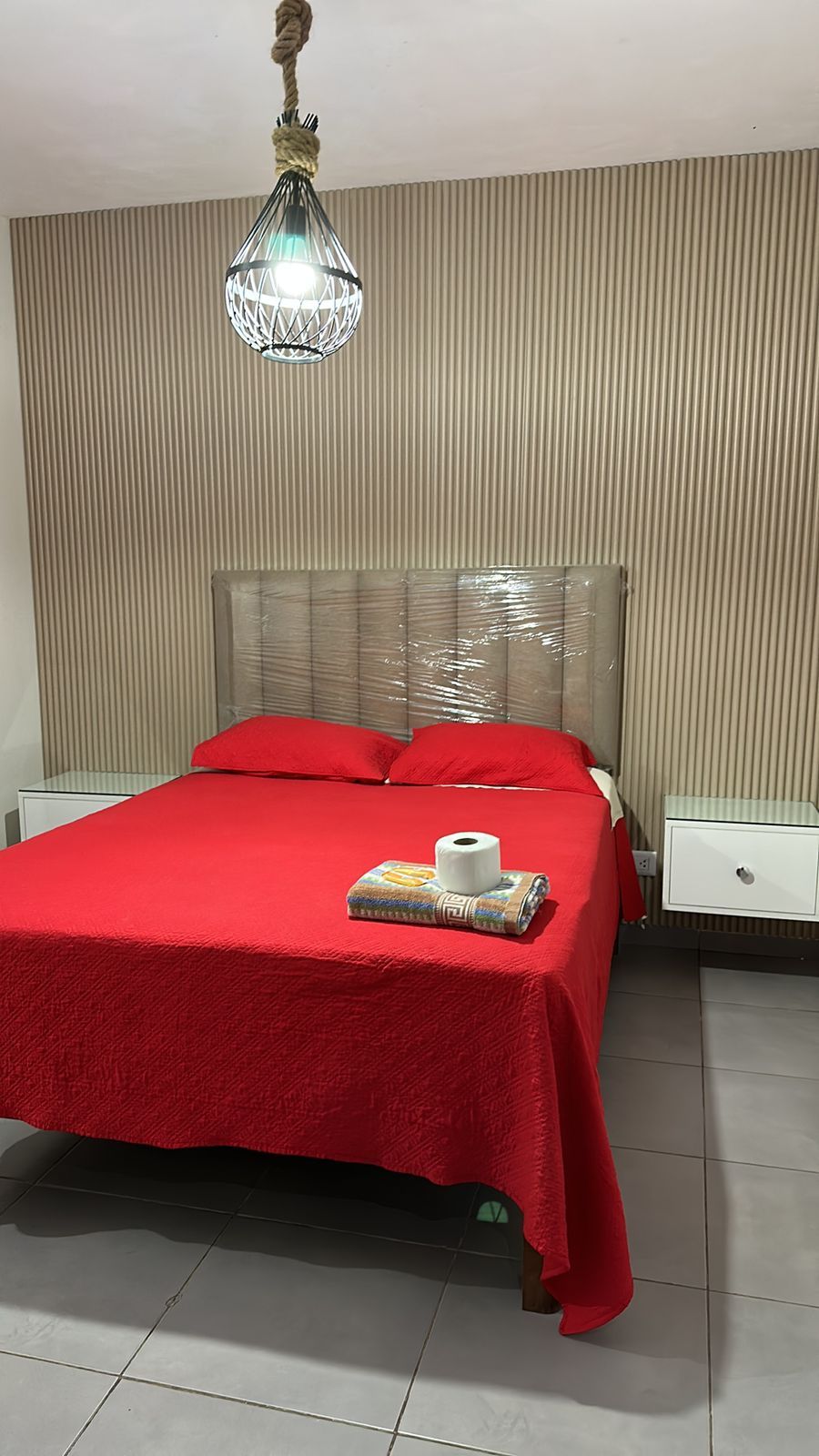 Un dormitorio con cama, mesita de noche, lámpara colgante y una pared con líneas verticales.