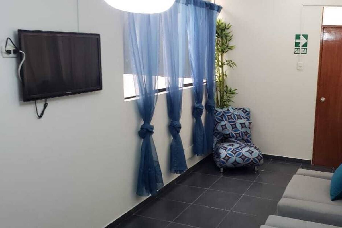 A reception area with a flat-screen TV mounted on the wall, a blue curtain, a patterned chair, a sofa with a blue cushion, and a wooden door with a sign above it.