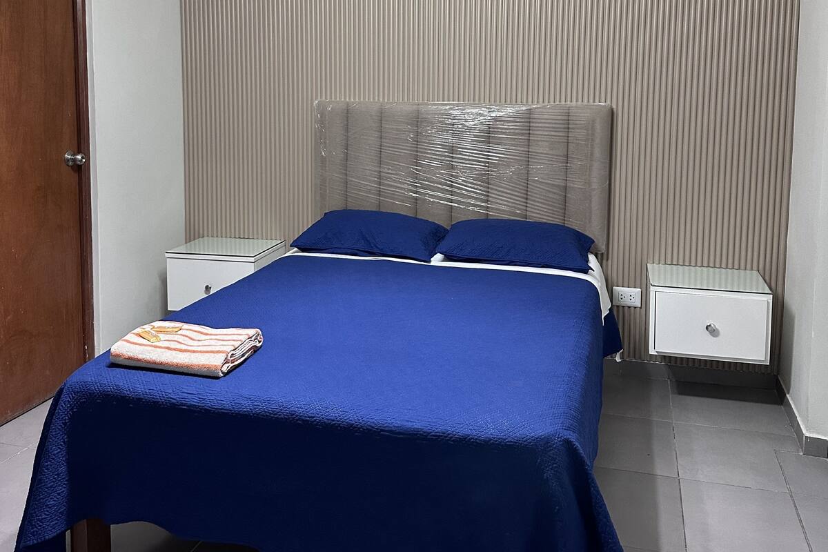 A bedroom with a bed, two bedside tables, a nightstand, a hanging light fixture, and a door.