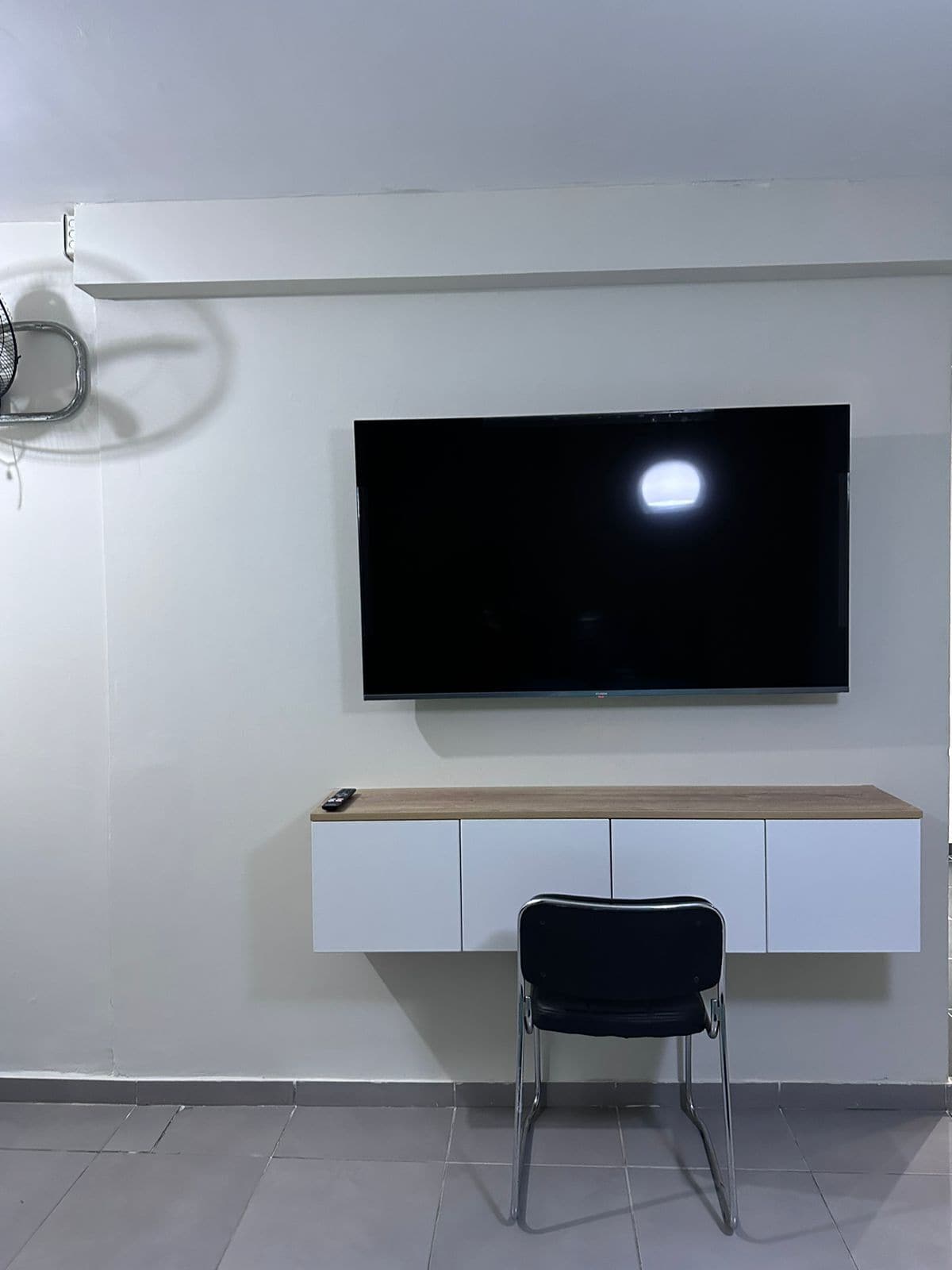 A modern living room with a flat-screen TV mounted on a white wall, a wooden bench with a remote, and a black chair.