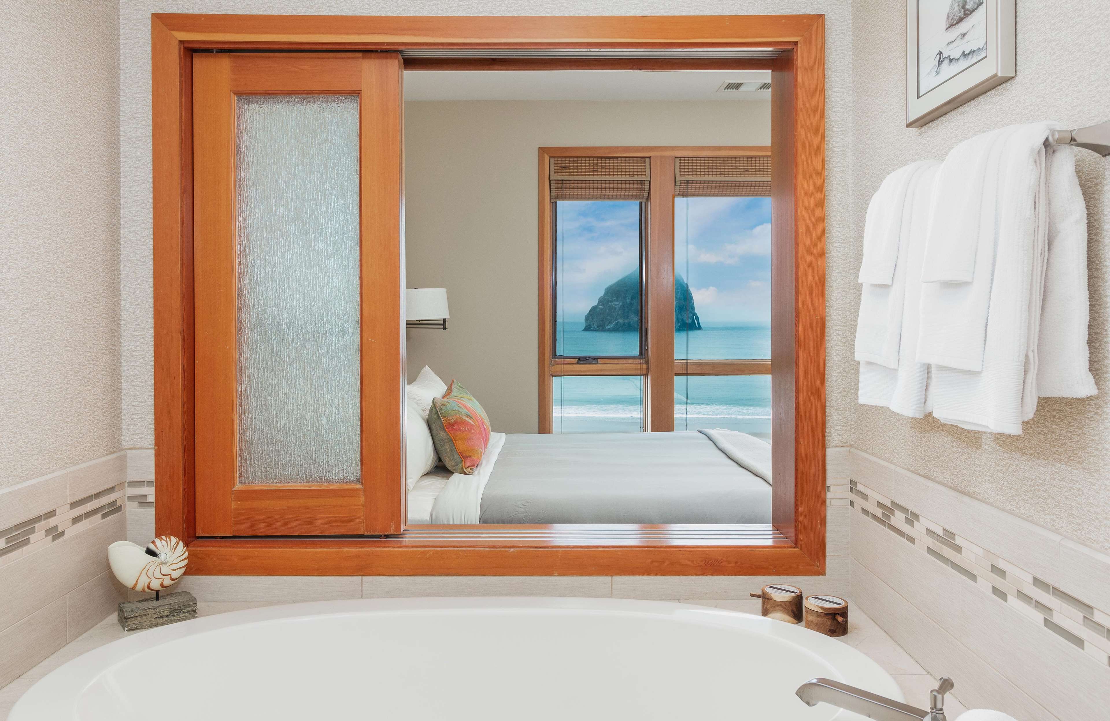 A bathroom with a large bathtub, a view of the ocean through a glass door, and a bed with pillows.
