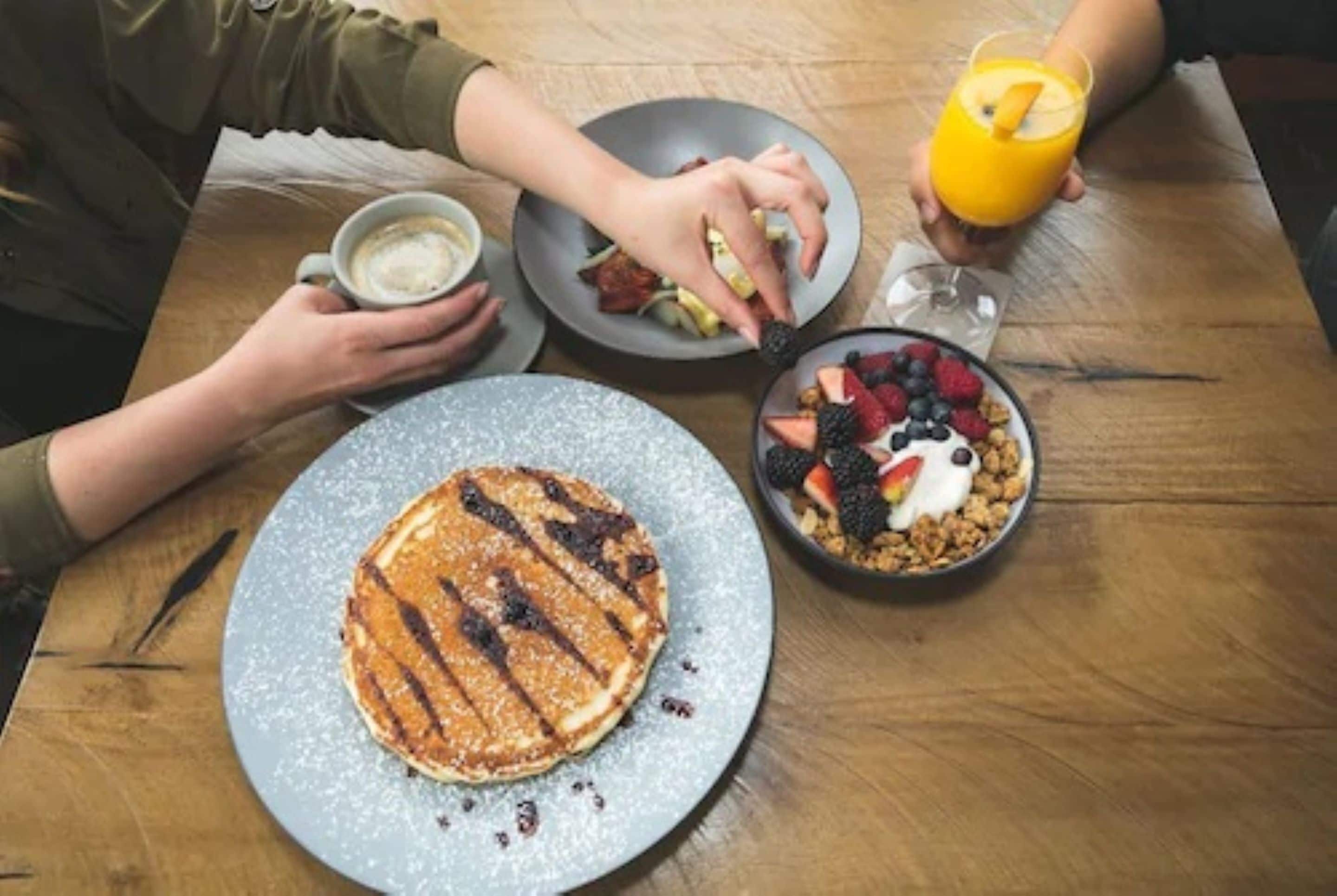Een ontbijttafel met pannenkoeken, een fruitschaal en een glas sinaasappelsap.