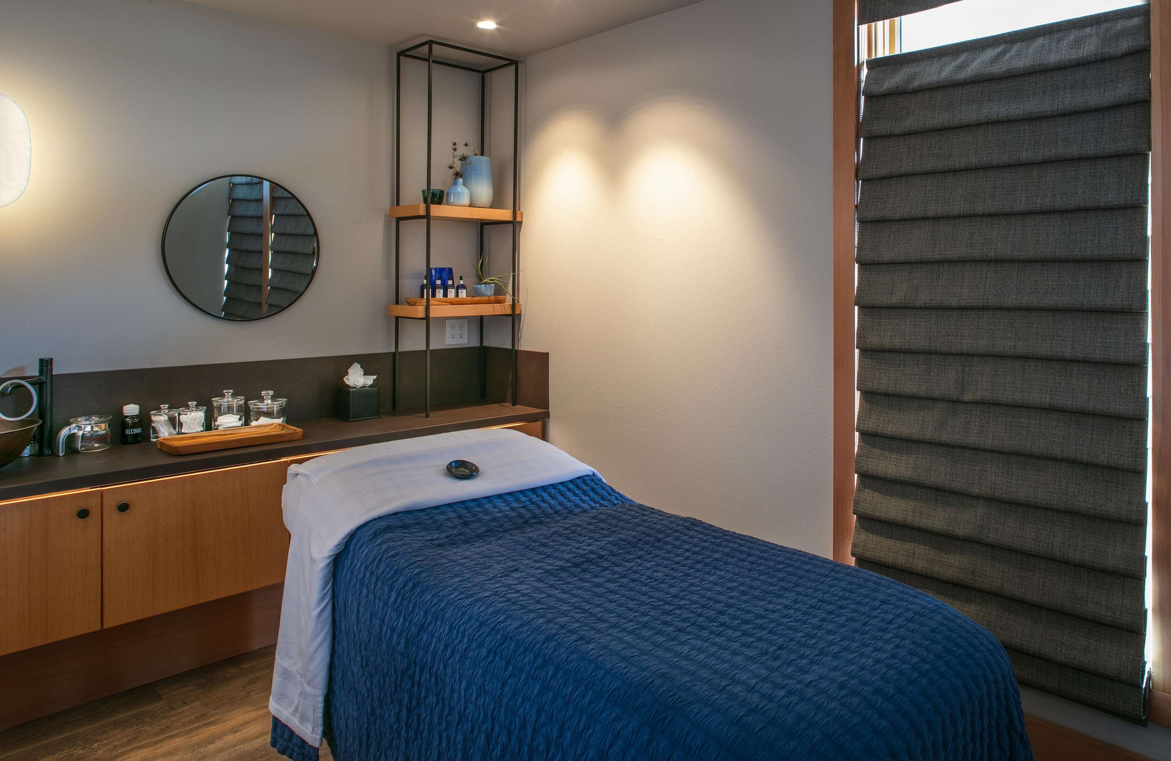 A bedroom with a bed, a bedside table with a lamp, a mirror, a shelf with toiletries, and a window with blinds.