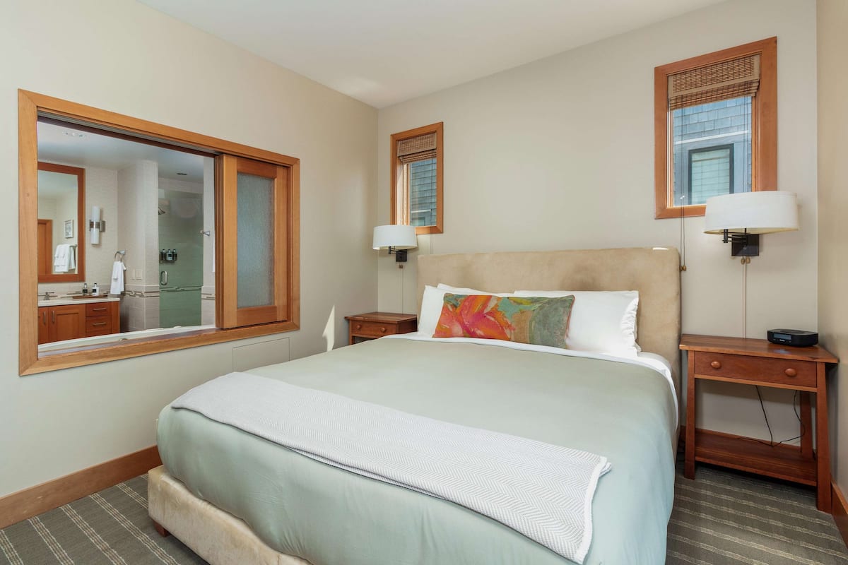 Une chambre d’hôtel avec un grand lit, deux tables de chevet, une commode, un miroir et une fenêtre avec des stores.