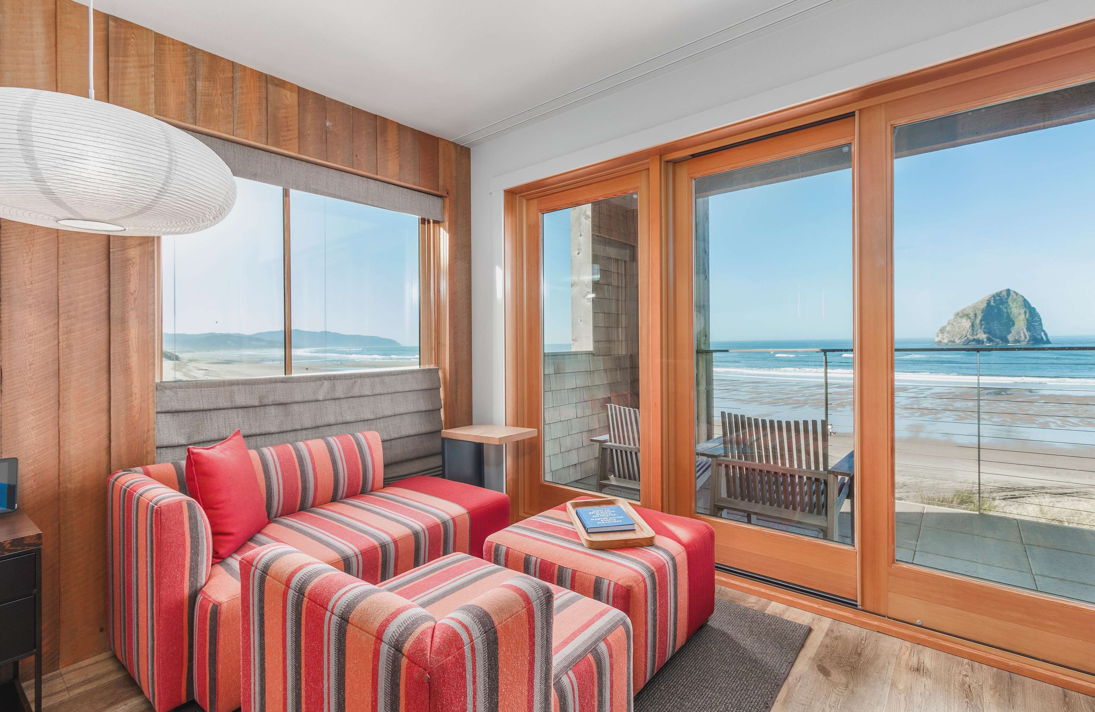 Ein modernes Zimmer mit einer großen Schiebeglaswand, die auf einen Strand blickt, einem gestreiften Sofa und einem kleinen Tisch mit einem Buch.