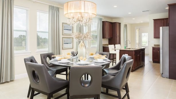 A dining area with a round table, chairs, and a chandelier. There is a kitchen in the background with cabinets and a refrigerator.