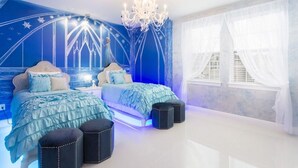 A bedroom with a bed, two ottomans, a chandelier, and a window with sheer curtains.