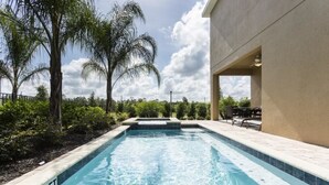 A swimming pool with a 5 ft depth marker, surrounded by palm trees and a patio area with outdoor furniture.