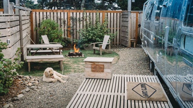 Een achtertuin met een vuurkuil, twee houten stoelen, een hond en een houten terras dat naar een busje leidt.