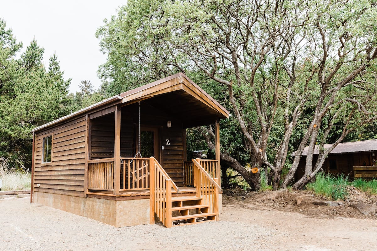 Una cabaña de madera con un porche y una sola puerta, rodeada de árboles.