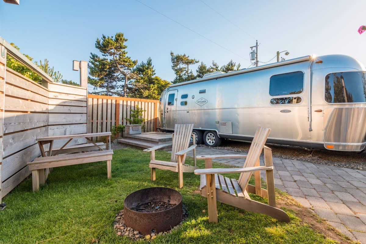 Una caravana estacionada en un patio trasero con un brasero, sillas de madera y una terraza de madera.