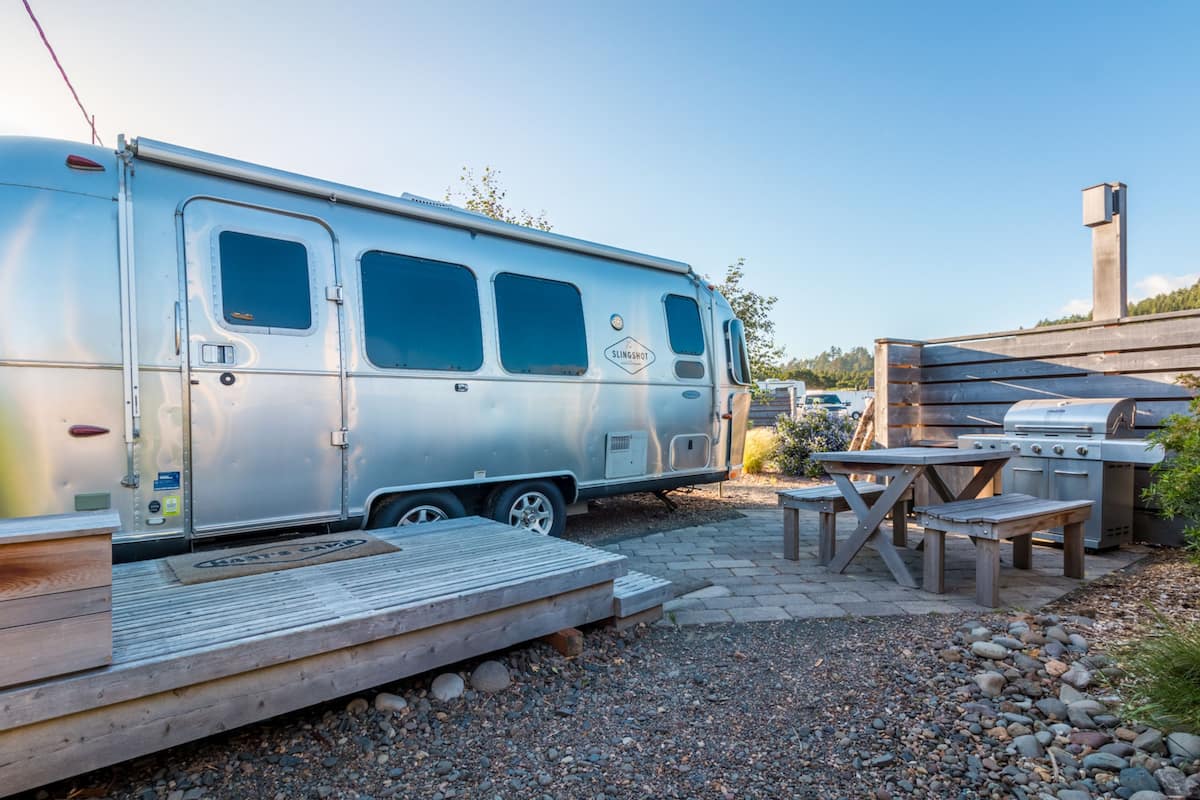 Una caravana plateada con puerta corredera, mesa de picnic y zona de barbacoa.