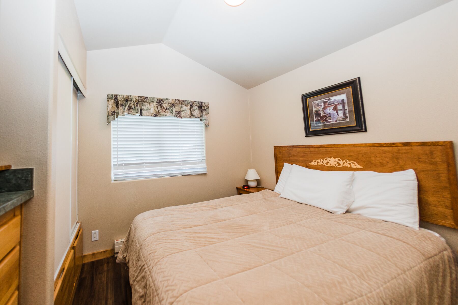 A bedroom with a bed, a nightstand, a lamp, a window with blinds, and a framed picture on the wall.