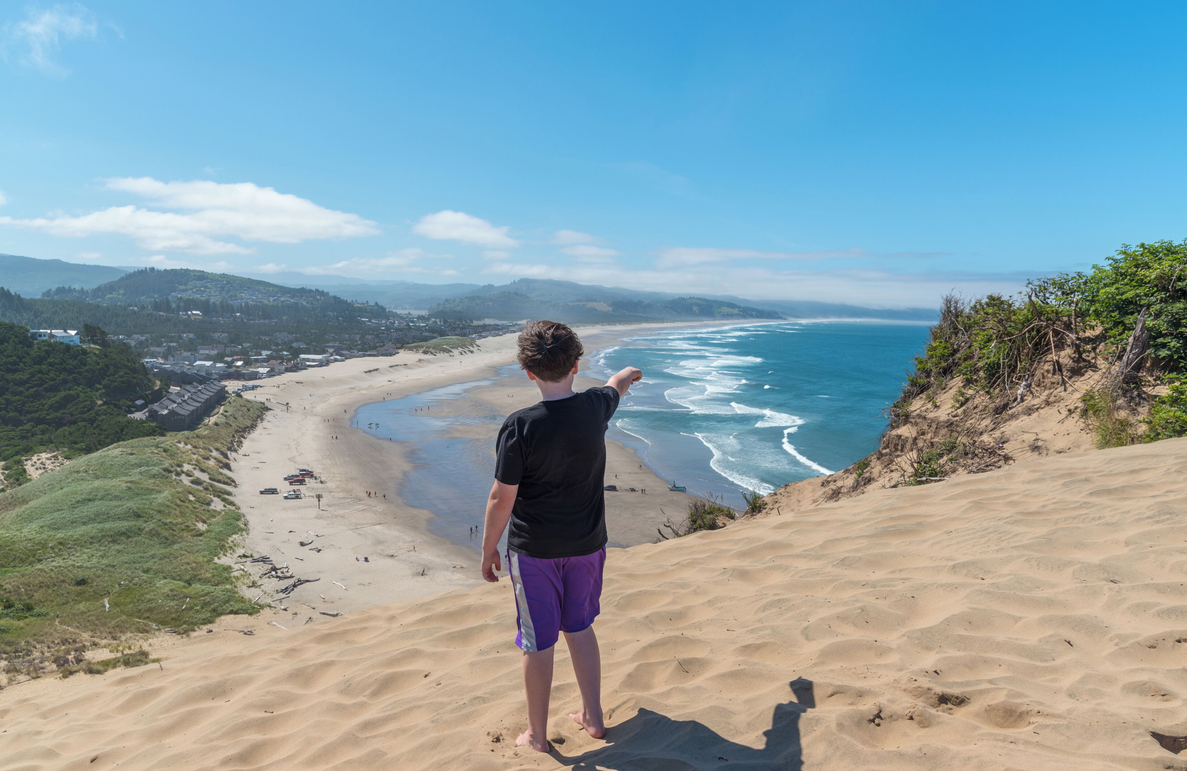 A person standing on a sandy slope overlooking a beach with waves, with a town in the background.