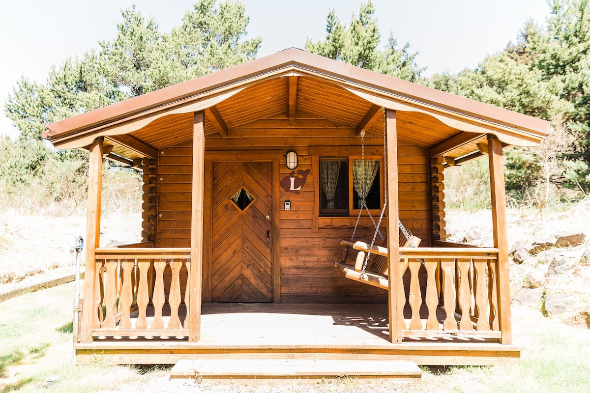 Una cabaña de madera con un porche, una hamaca y una puerta con un ventanal en forma de rombo.