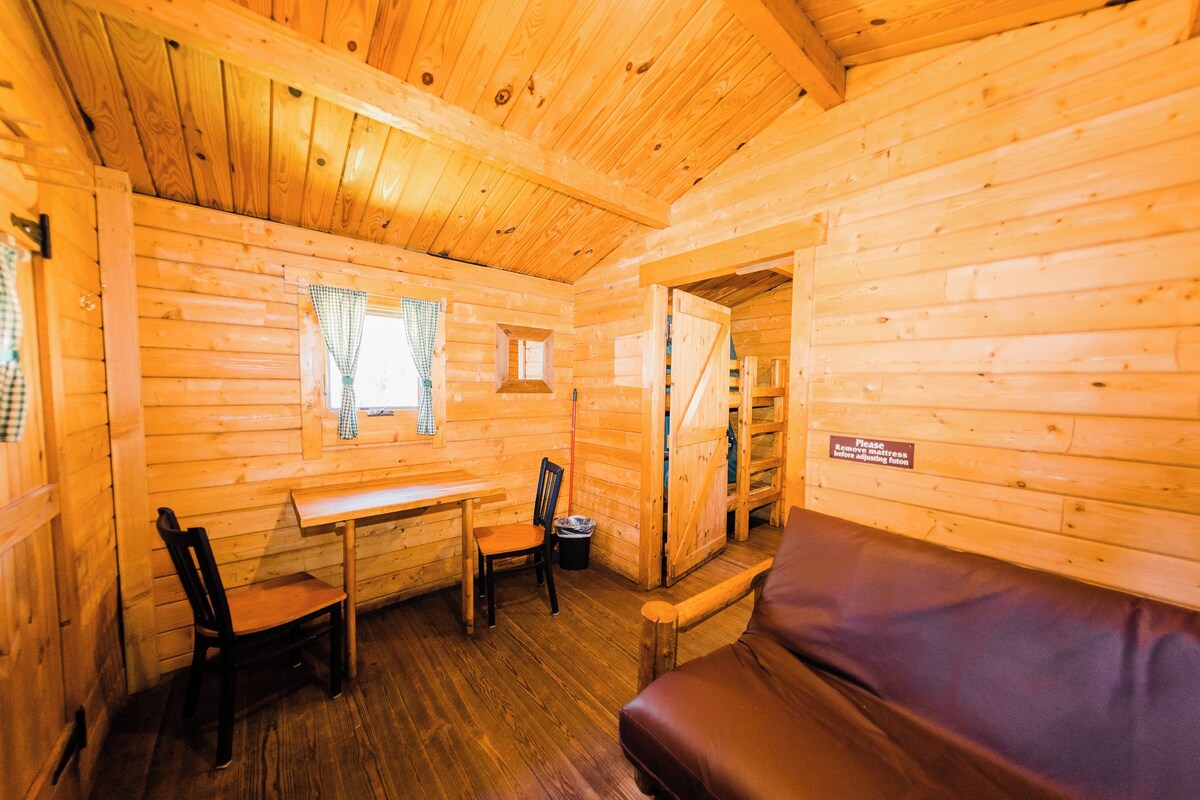 Une pièce chaleureuse aménagée dans un chalet en rondins, avec un intérieur en bois, une table à manger entourée de chaises, un canapé et une petite fenêtre agrémentée de rideaux.