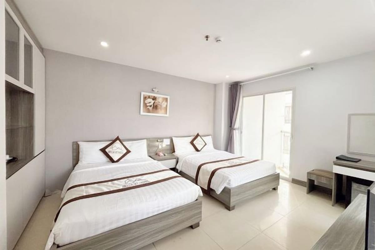 A hotel room with two beds, a desk, a chair, a window with curtains, and a framed picture on the wall.