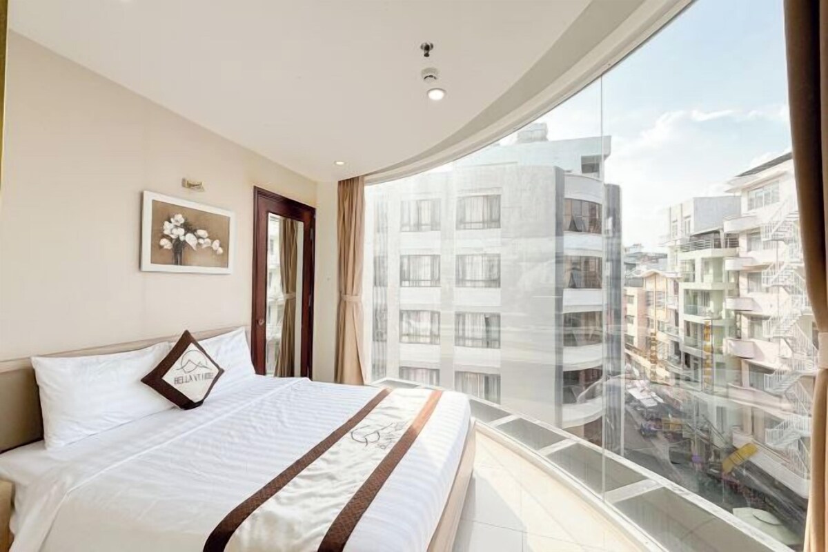 A bedroom with a bed, a window, a framed picture, and a view of buildings.