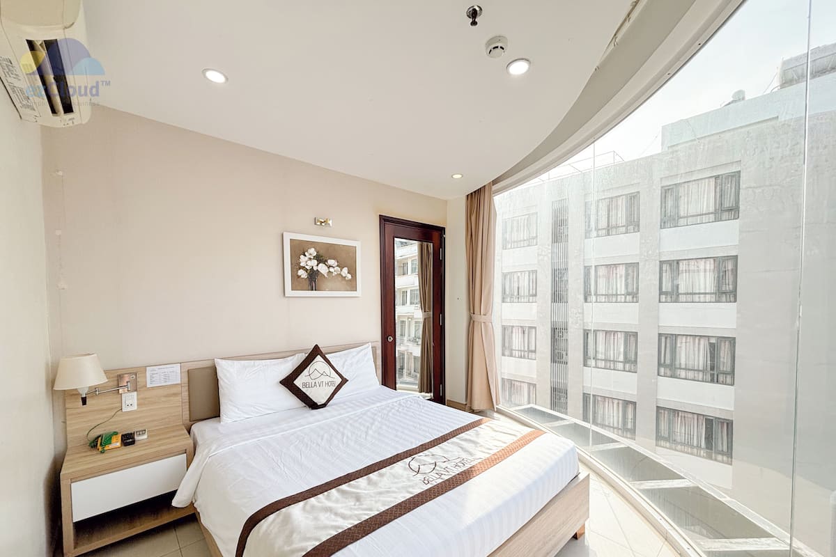 A modern hotel room with a large bed, a bedside table with a lamp, a small desk, and a view of the city through large windows.