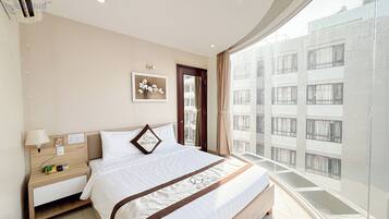 A modern hotel room with a large bed, a bedside table with a lamp, a small desk, and a view of the city through large windows.