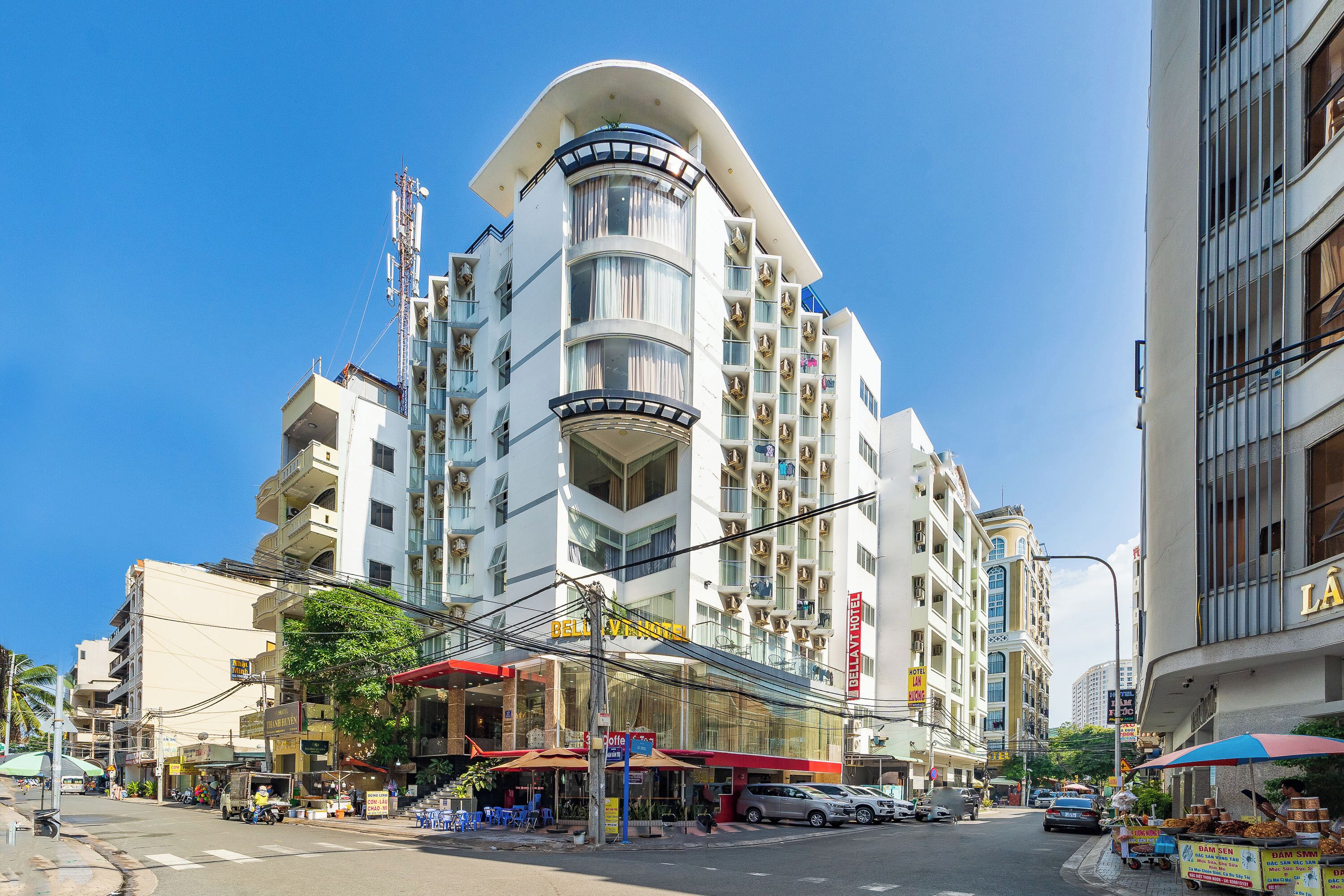A multi-story hotel with a curved corner design, balconies, and a ground-level commercial area with outdoor seating.