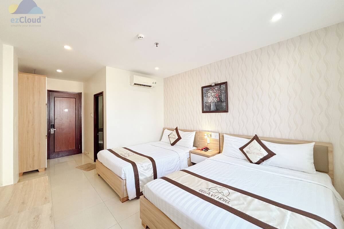 A hotel room with two beds, a wooden wardrobe, an air conditioning unit, a framed artwork on the wall, and a small bedside table with a lamp.