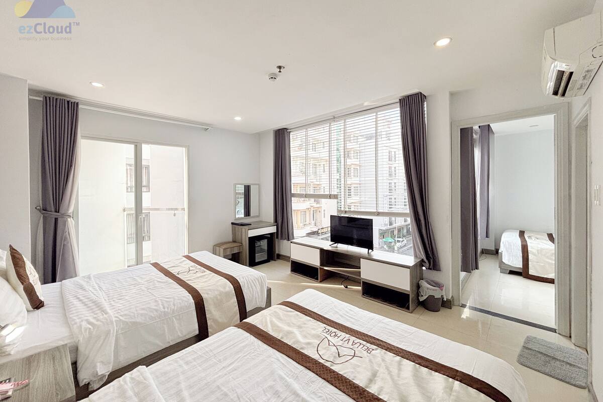 A hotel room with two beds, a desk, a TV, and a large window with curtains.
