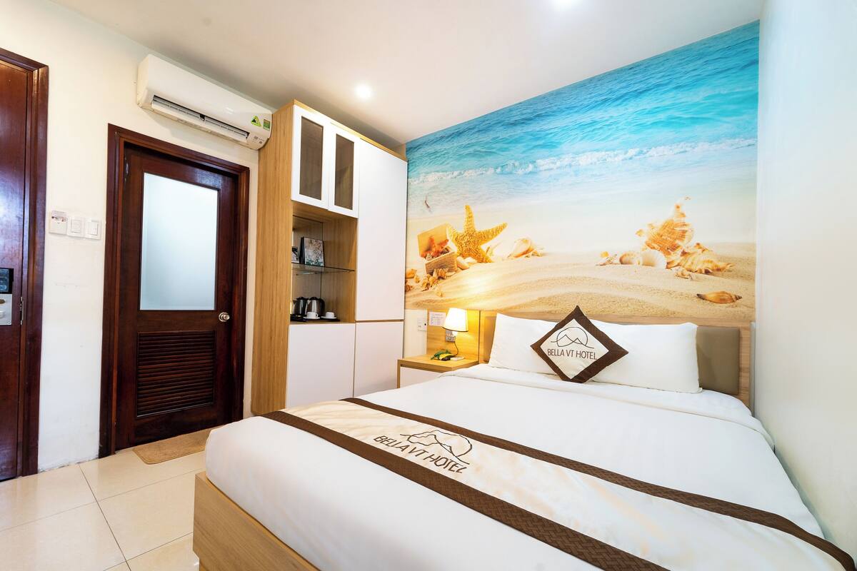 A hotel room with a bed, a nightstand, a lamp, a wall-mounted air conditioner, and a beach-themed mural.
