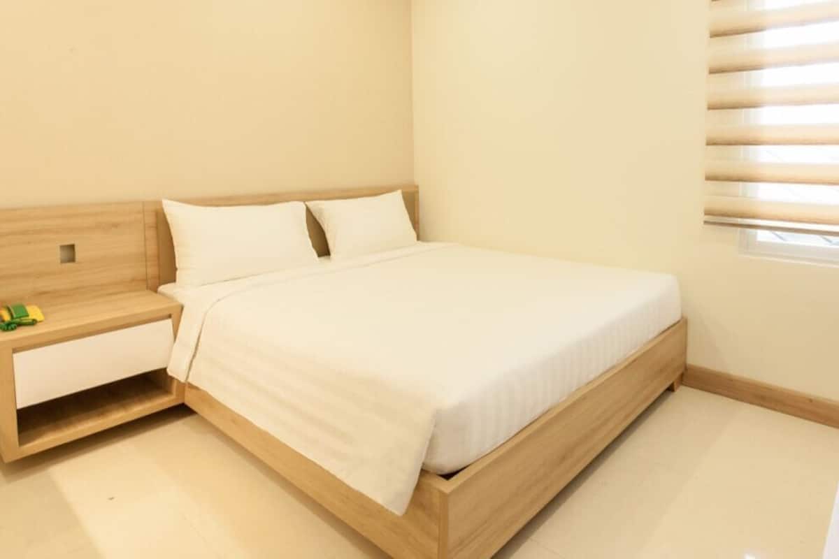 A bedroom with a bed, a nightstand, a wooden headboard, and a window with blinds.