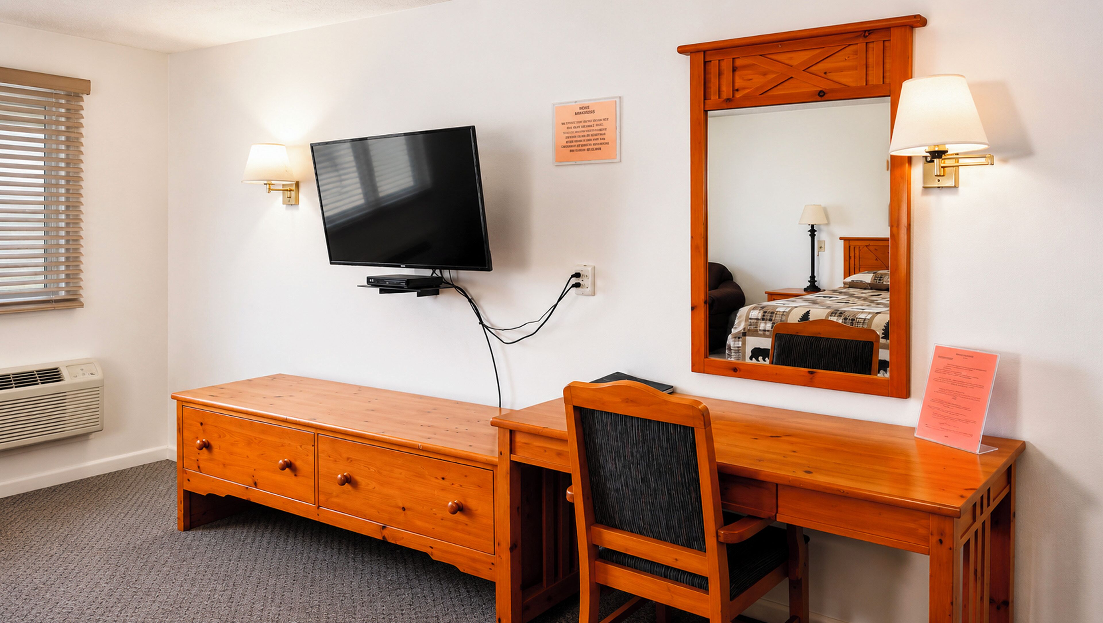 Een hotelkamer met een houten dressoir, een aan de muur bevestigde televisie, een spiegel met lamp en een bed met een geruit sprei.