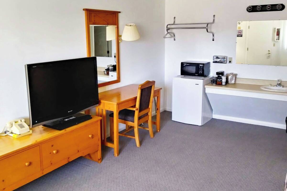 A compact hotel room with a television, wooden desk, chair, microwave, sink, and a mirror.