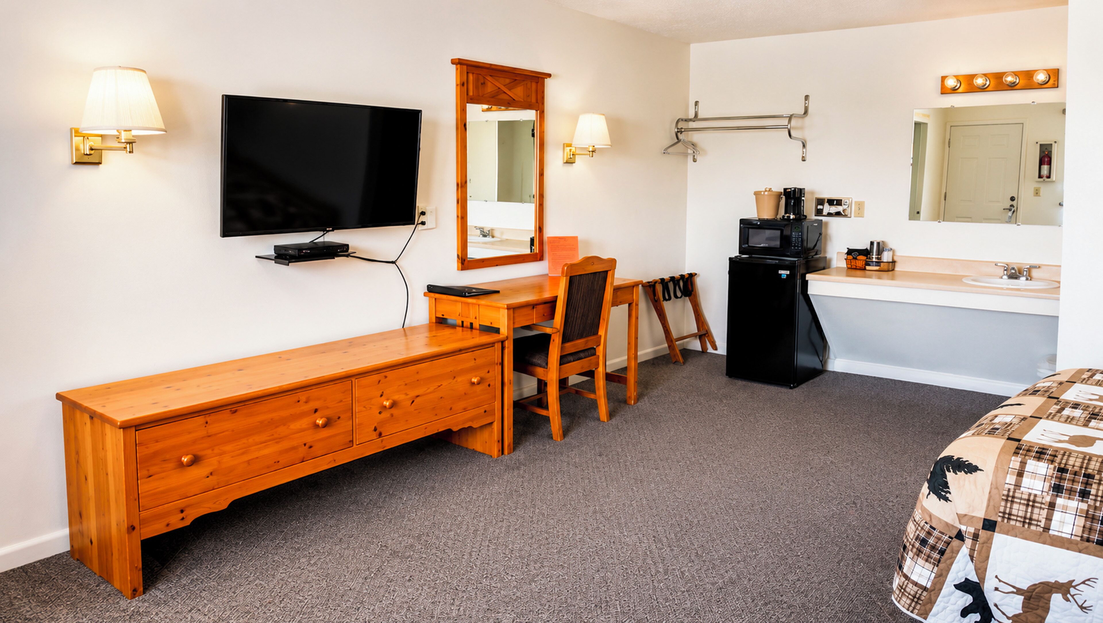 A hotel room with a wooden dresser, a desk with a chair, a television, a mirror, a refrigerator, a sink, and a bed with a plaid bedspread.