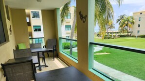 A balcony with a table and chairs, a view of a well-maintained lawn and palm trees, and a building with multiple windows.