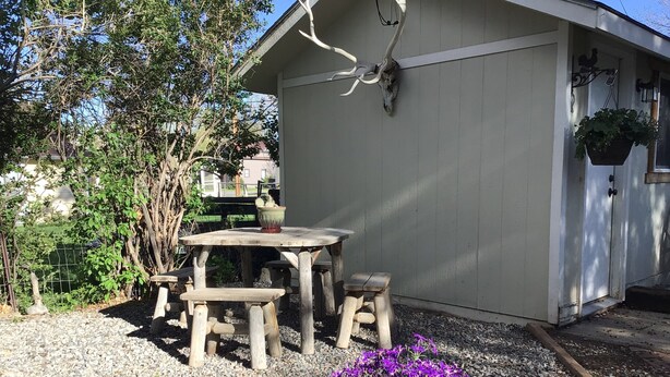 Una mesa de madera y bancos en un jardín con suelo de grava, un edificio blanco con puerta de garaje y una planta en maceta con flores púrpuras.