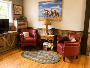 A cozy living room with two leather armchairs, a wooden coffee table, a lamp, a painting of horses, and a bookshelf. - “The Wyoming,” A Beautiful historic downtown home. (Cody)