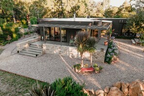 Une maison moderne dotée d’un patio couvert, d’un escalier en pierre et d’un jardin où poussent diverses plantes et fleurs.