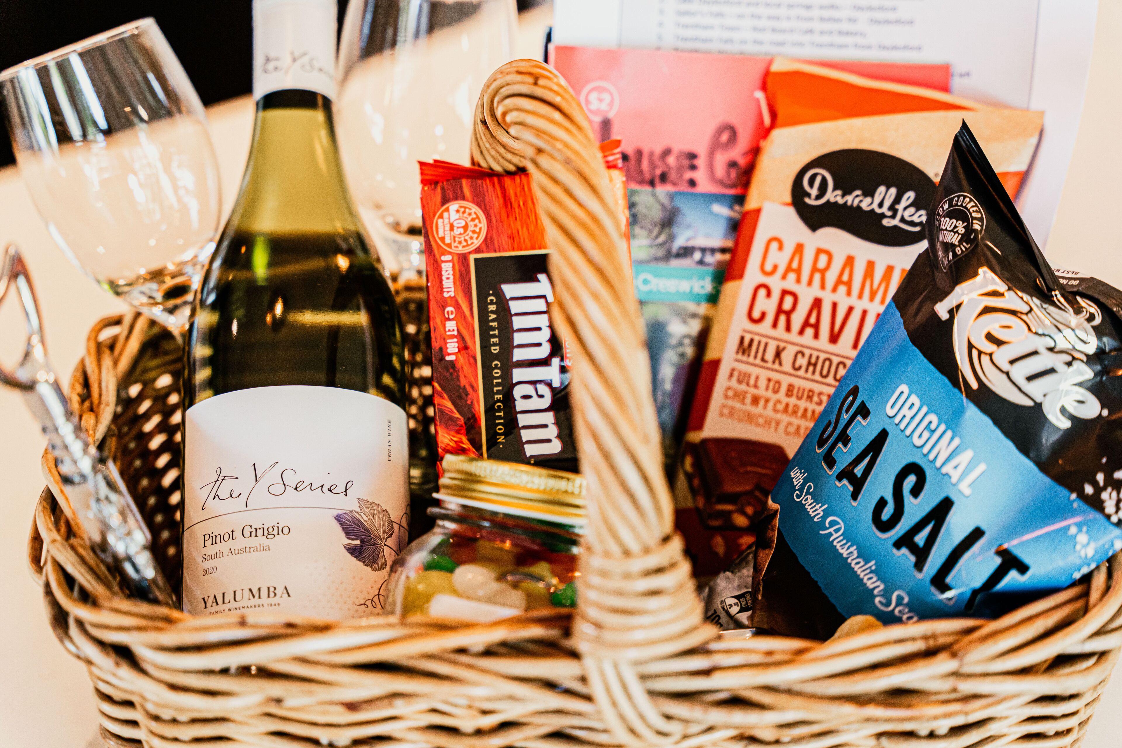 A basket containing a bottle of Pinot Grigio wine, a jar of Tim Tam chocolate, a Tim Tam bar, a bag of Caramel Cravings, and a bag of Sea Salt.