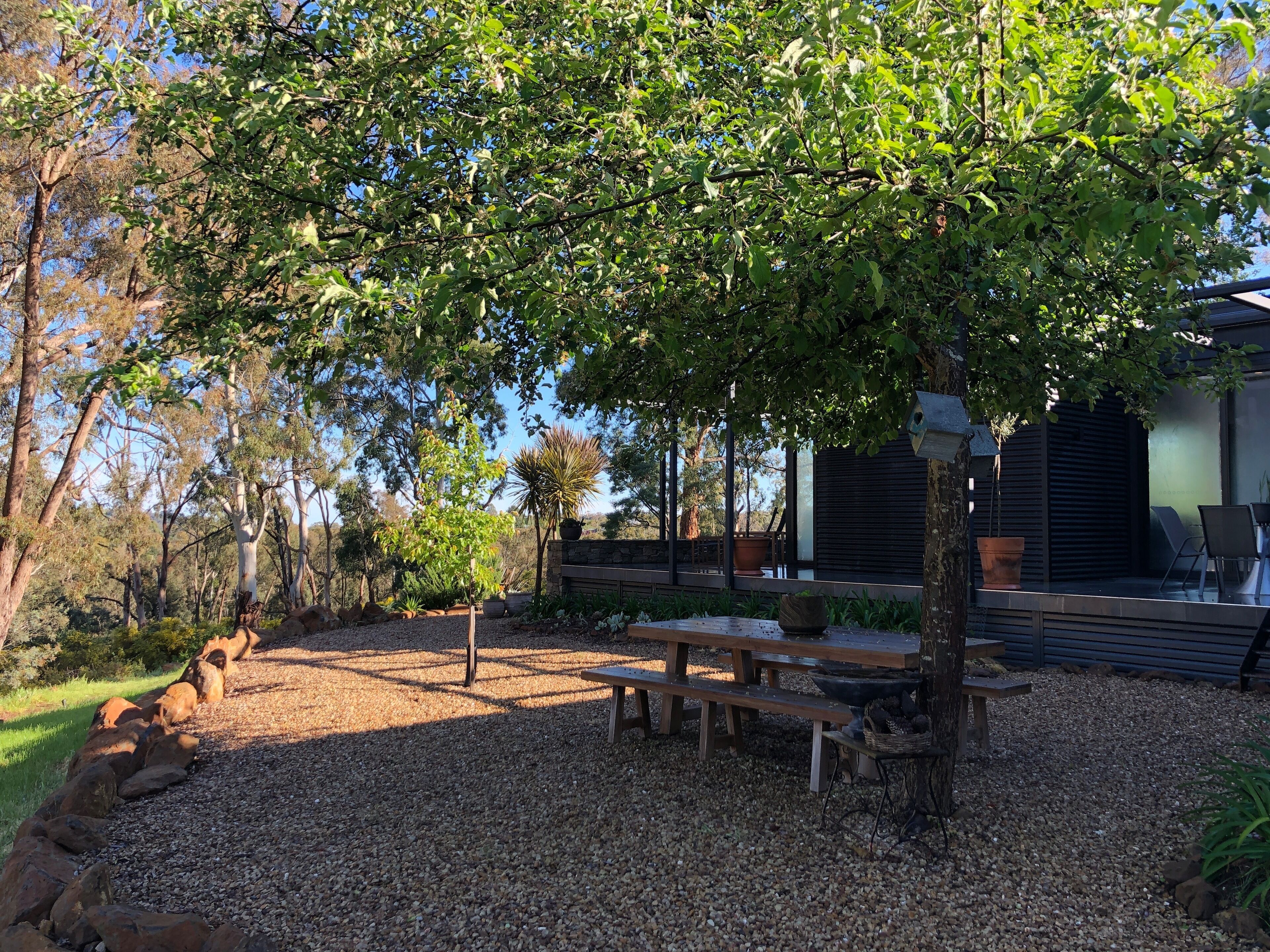 A patio area with a wooden table and benches, surrounded by gravel and rocks, with trees and a building in the background.