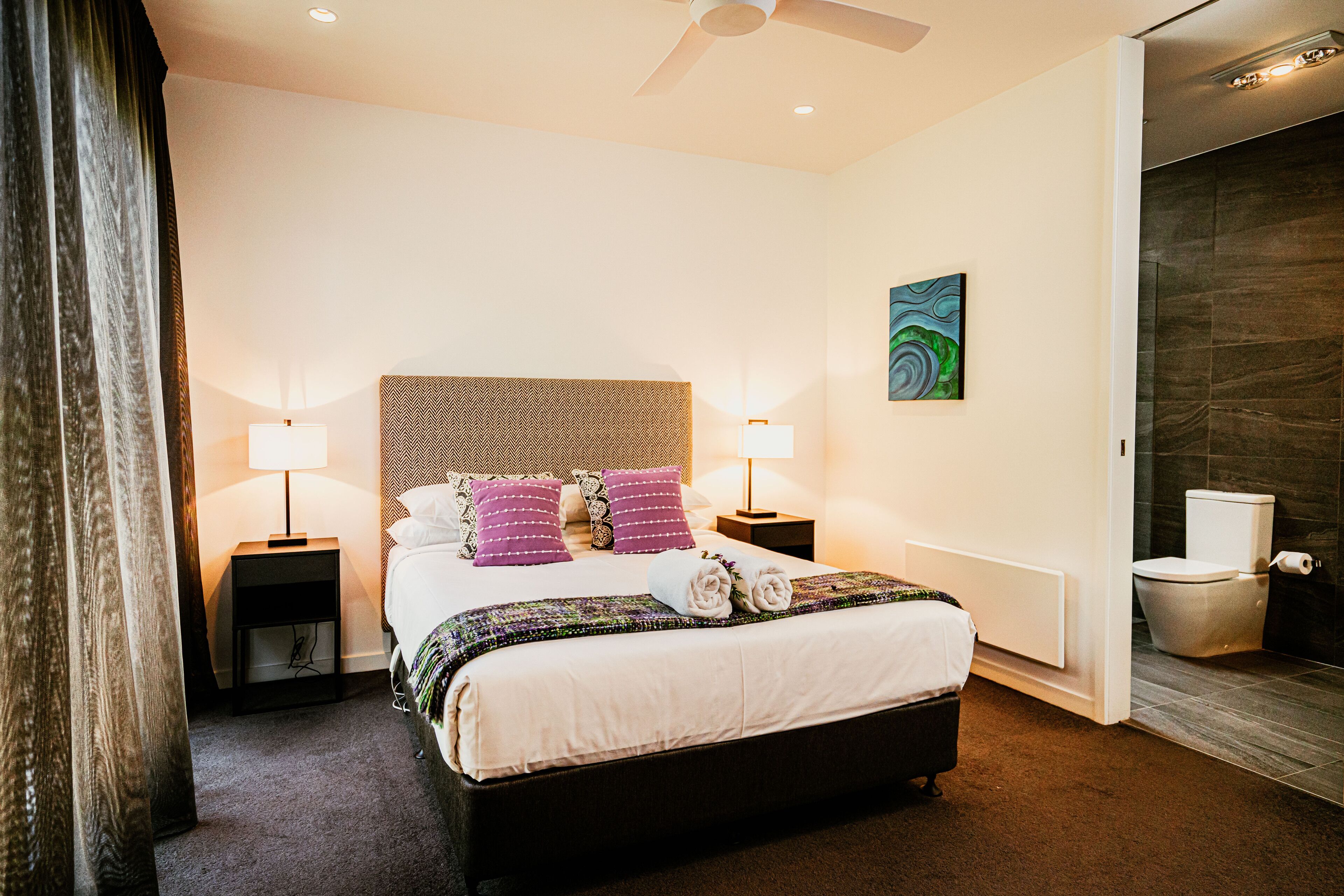 Une chambre d’hôtel avec un grand lit, deux tables de chevet avec des lampes, un ventilateur de plafond, un tableau au mur et une salle de bain avec des toilettes et une douche.