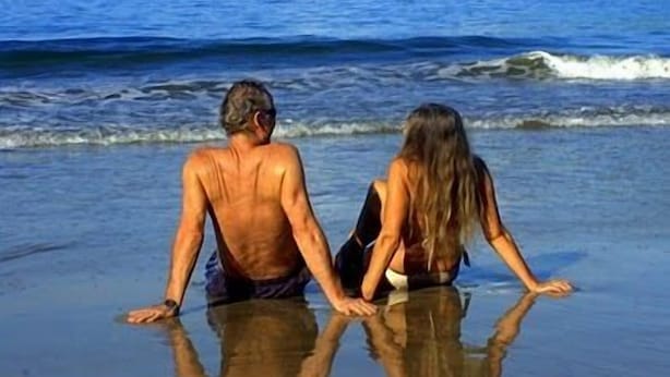 Zwei Personen sitzen am Strand und blicken auf das Meer, während Wellen rollen.