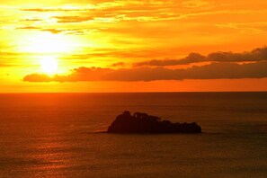 Un coucher de soleil sur une mer calme, avec la silhouette d’une formation rocheuse au premier plan.