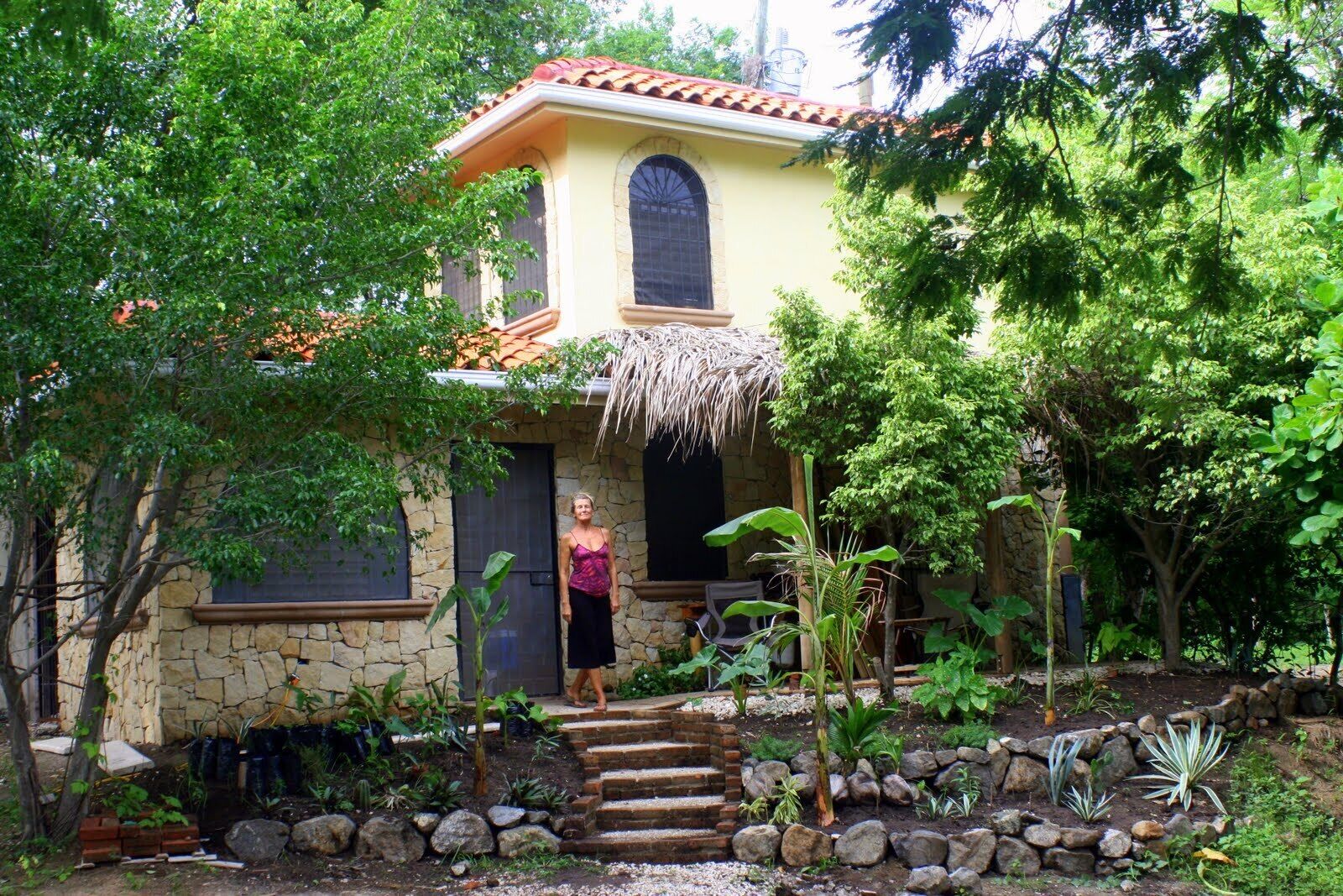 Ein Haus mit Natursteinfassade, einem Reetdach-Vordach und einem Garten mit verschiedenen Pflanzen sowie einem Steinweg, der zur Tür führt.