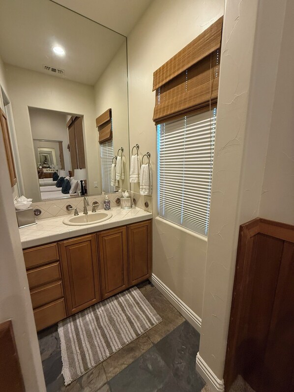 A modern bathroom with a large mirror, wooden cabinets, a sink, and a window with blinds.