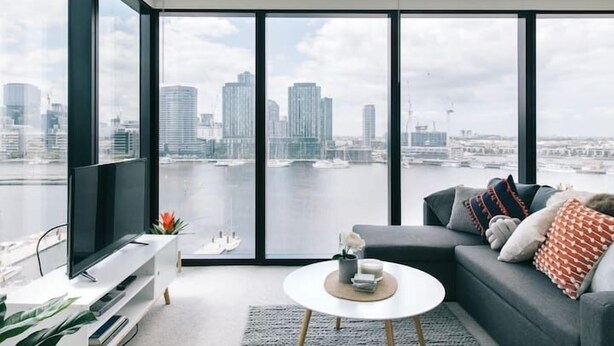 Een moderne woonkamer met een groot raam, een grijze bank, een witte salontafel en een flatscreen televisie.
